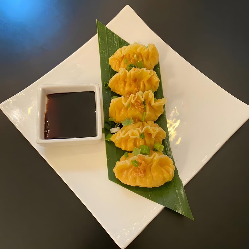 Five golden Thai dumplings on a banana leaf with a side of soy sauce on a white plate.