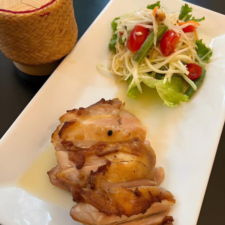 Grilled chicken with papaya salad and a woven sticky rice container on a white plate.