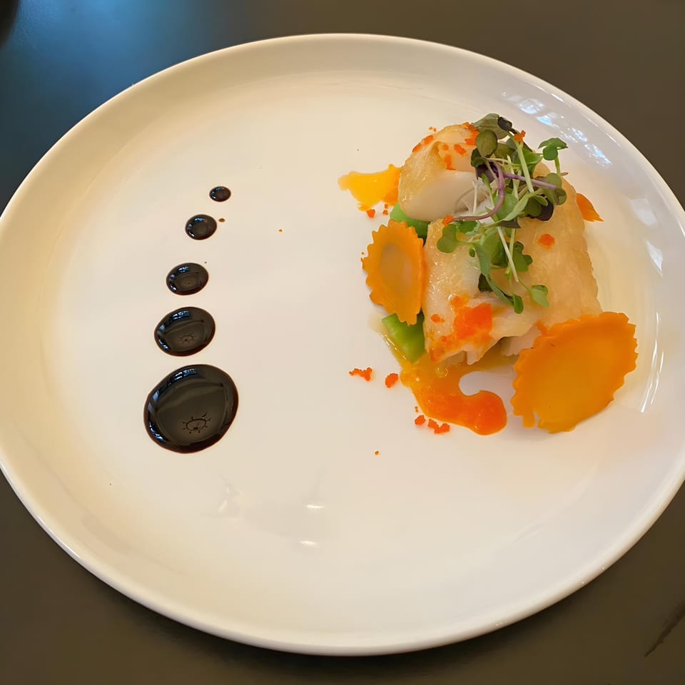 Thai Den's elegant dish with fish, microgreens, orange sauce, and dark balsamic dots on a white plate.
